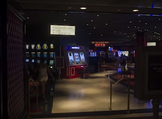 A dimly lit casino with several rows of slot machines, some displaying bright graphics and lights. There are a few people scattered throughout the area, engaging with the machines. The floor is reflective and polished, adding to the ambiance, while the ceiling is dotted with numerous small lights.