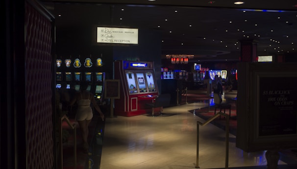 A dimly lit casino with several rows of slot machines, some displaying bright graphics and lights. There are a few people scattered throughout the area, engaging with the machines. The floor is reflective and polished, adding to the ambiance, while the ceiling is dotted with numerous small lights.