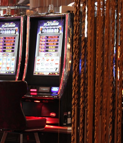 A pair of casino slot machines, each with detailed screens displaying various game outcomes and jackpot amounts. The chairs in front of the slot machines are dark and cushioned for comfort. The background features a hanging curtain made of golden ropes and other casino decorations are visible.
