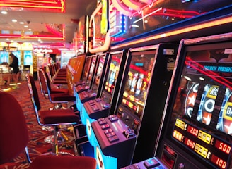 A brightly lit casino interior featuring a row of colorful slot machines. The neon lights create a vibrant atmosphere, with patterned carpeting adding to the decor. Stools are aligned neatly in front of each machine.