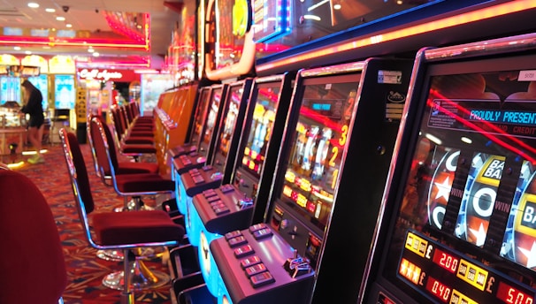 A brightly lit casino interior featuring a row of colorful slot machines. The neon lights create a vibrant atmosphere, with patterned carpeting adding to the decor. Stools are aligned neatly in front of each machine.