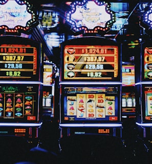 A row of colorful slot machines lit with bright neon lights, showcasing various graphical elements on the screens, indicating different games and jackpots. The atmosphere suggests a lively casino setting, bustling with excitement.