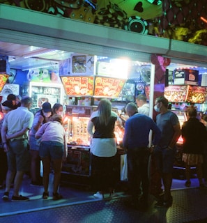 A group of people standing in front of an illuminated amusement arcade, engaging with various gaming machines. The environment is bustling with activity, and colorful lights create a vibrant atmosphere. The ceiling is decorated with large, playful images of dice and other game-related graphics, enhancing the lively mood.