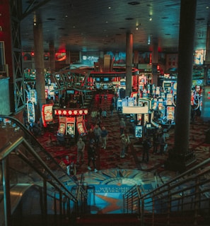 A bustling casino floor filled with groups of slot machines and people walking around. The atmosphere is vibrant with neon lights and illuminated signs. The interior has a ceiling supported by large pillars and there are stairs leading down to the gaming area.
